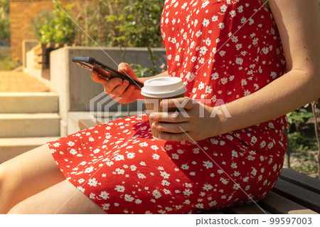 Young woman scrolling social media on mobile phone on bench in park. Young woman in casual clothes using mobile phone and holding cup of coffee while sitting in park on sunny day. Side view of Young woman scrolling social media on mobile phone on bench in park. Young woman in casual clothes using mobile phone and holding cup of coffee while sitting in park on sunny day. Side view of 99597003