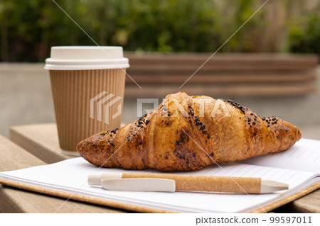 Fresh baked chocolate croissant on paper cup with lid for tea to go. Empty paper notebook. Freelancer's place of work. Study and work. Remote business education. Making plans for next week year. Being Fresh baked chocolate croissant on paper cup with lid for tea to go. Empty paper notebook. Freelancer's place of work. Study and work. Remote business education. Making plans for next week year. Being 99597011