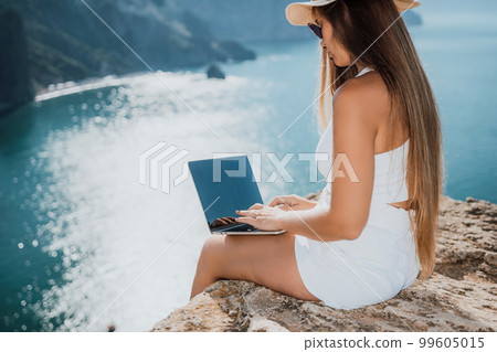 Digital nomad, woman in the hat, a business woman with a laptop sits on the rocks by the sea during sunset, makes a business transaction online from a distance. Freelance, remote work on vacation. 99605015
