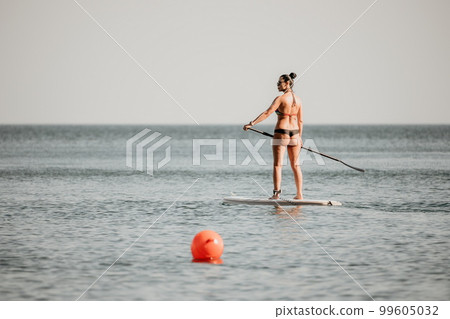 Sea woman sup. Silhouette of happy middle aged tanned woman in rainbow bikini, surfing on SUP board, confident paddling through water surface. Idyllic sunset. Active lifestyle at sea or river. 99605032