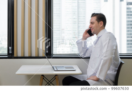 Young caucasian doctor in white gown use mobile phone while checking email on laptop computer. Sitting near the window in cafeteria. Outside the window is a city view with high rise buildings. Young caucasian doctor in white gown use mobile phone while checking email on laptop computer. Sitting near the window in cafeteria. Outside the window is a city view with high rise buildings. 99607630