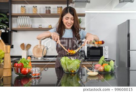 Young asian woman dressed in an apron mix the vegetables in a salad bowl together with a wooden ladle. Tomato, bell pepper and lettuce. Morning atmosphere in a kitchen. 99608015