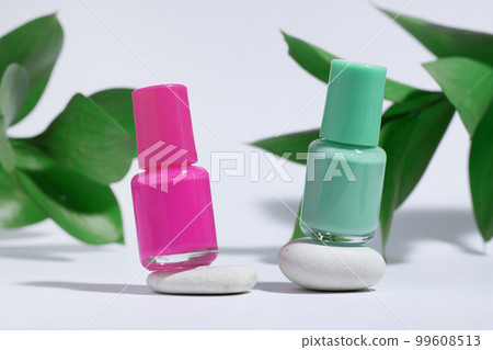 Manicure or pedicure background. Creative mockup of isolated cosmetics bottles with pink and green nail polish with plant, on stone, on white background with hard shadows from plants. Manicure or pedicure background. Creative mockup of isolated cosmetics bottles with pink and green nail polish with plant, on stone, on white background with hard shadows from plants. 99608513