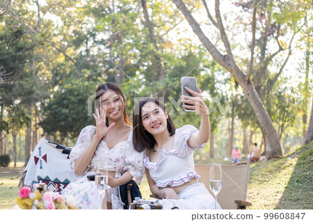 Two beautiful young Asian women picnic in the green park together, taking selfies with their phone they are having picnic,they talk happily 99608847