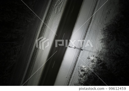 An atmospheric drone photo of a winter road winding through a snow-covered forest at night. The trees are adorned with a delicate layer of hoarfrost, creating a magical winter wonderland 99612459