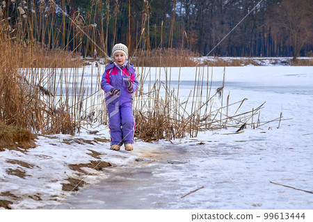 A cute child in a lilac jumpsuit walks on the lake in a winter park 99613444