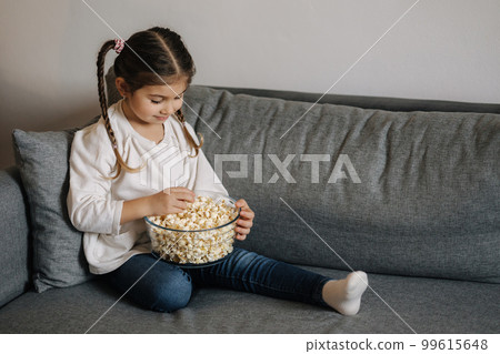 Adorable little girl sits on sofa and watching TV at home. Cute girl eating popcorn. Holiday mood 99615648