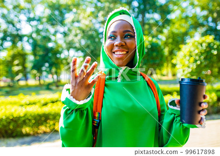 modern afro woman in green hijab with bright make up and piercing nose drinking coffee outdoor and communicating with her friend modern afro woman in green hijab with bright make up and piercing nose drinking coffee outdoor and communicating with her friend 99617898