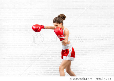Attractive caucasian young woman in red boxing gloves and boxer shorts practice a punch on loft white background. 99618773