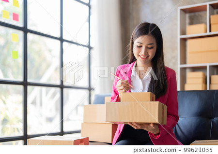 Young woman, an Asian online business owner, smiles, prepares parcel boxes and checks online orders for products to be delivered to customers Young woman, an Asian online business owner, smiles, prepares parcel boxes and checks online orders for products to be delivered to customers 99621035
