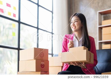 Young woman, an Asian online business owner, smiles, prepares parcel boxes and checks online orders for products to be delivered to customers Young woman, an Asian online business owner, smiles, prepares parcel boxes and checks online orders for products to be delivered to customers 99621039