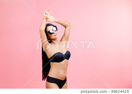 Young woman in summer black swimsuit and sunglasses standing on pink background in studio 99624429