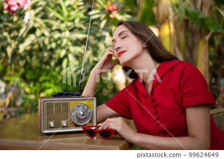 a beautiful girl in a red retro style dress listens to an antique radio. a beautiful girl in a red retro style dress listens to an antique radio. 99624649