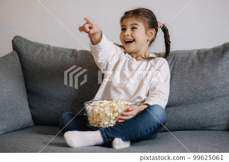 Adorable girl watching a cartoon emotionally and points his hand in the direction of the TV. little girl eating popcorn 99625061