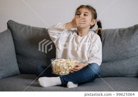 Little girl watching a cartoon emotionally and covers her mouth with hand in surprise. Popcorn. Holiday mood Little girl watching a cartoon emotionally and covers her mouth with hand in surprise. Popcorn. Holiday mood 99625064