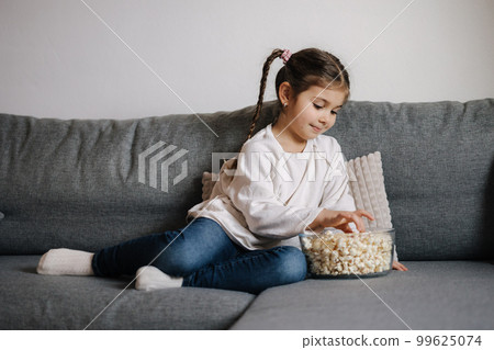 Adorable little girl sits on sofa and watching TV at home. Cute girl eating popcorn. Holiday mood Adorable little girl sits on sofa and watching TV at home. Cute girl eating popcorn. Holiday mood 99625074