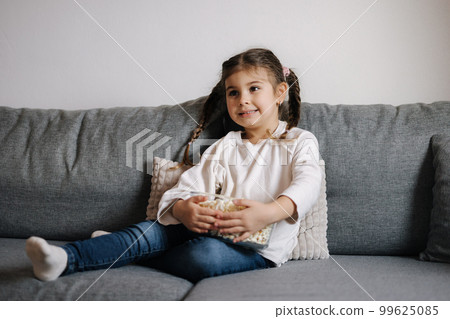 Adorable little girl sits on sofa and watching TV at home. Cute girl eating popcorn. Holiday mood 99625085