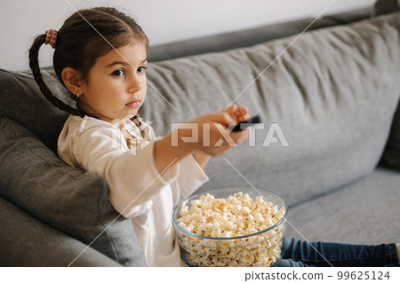 Cute little girl using remote controller of TV and watching cartoons. Four year girl eating popcorn on sofa 99625124