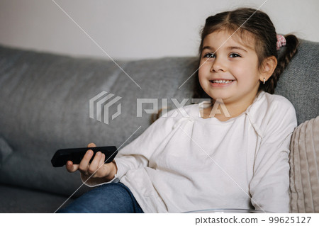 Portrait of cute little girl using remote controller of TV and watching cartoons. Small but already so grown up 99625127