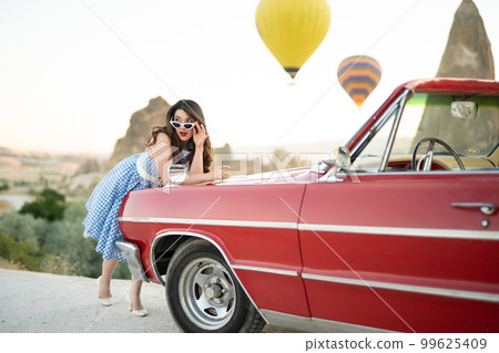 beautiful girl in retro style posing near a vintage red cabriolet car on background of balloons in Cappadocia. 99625409