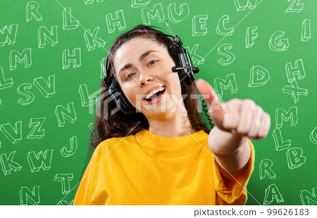Language learning and listening.Portrait of a young smiling woman wearing headphones, giving a thumbs up.Green background with letters.The concept of learning foreign languages Language learning and listening.Portrait of a young smiling woman wearing headphones, giving a thumbs up.Green background with letters.The concept of learning foreign languages 99626183