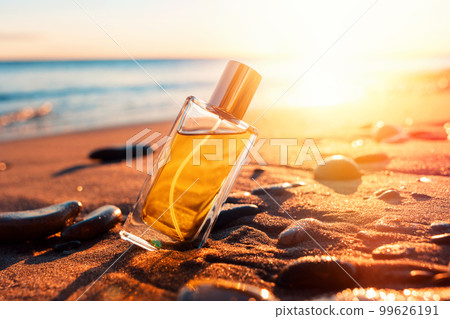 A transparent glass perfume bottle with a golden lid on a sandy beach. Close-up, view from the lower angle. Sunset in the background. Copy space. Perfume advertising concept 99626191