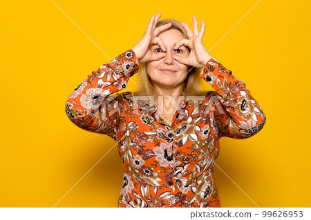 Portrait of astonished caucasian blonde woman spying, seeing amazing shocking event, looking through fingers imitating binoculars, wearing patterned dress. Isolated on yellow background Portrait of astonished caucasian blonde woman spying, seeing amazing shocking event, looking through fingers imitating binoculars, wearing patterned dress. Isolated on yellow background 99626953