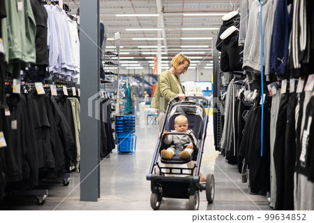 Casualy dressed mother choosing sporty shoes and clothes products in sports department of supermarket store with her infant baby boy child in stroller 99634852
