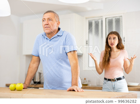 Adult daughter scolds an elderly father while he is preparing dinner in kitchen Adult daughter scolds an elderly father while he is preparing dinner in kitchen 99636282