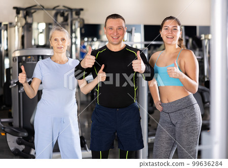 Two sportswomen and one sportsman standing in middle of gym showing thumbs up Two sportswomen and one sportsman standing in middle of gym showing thumbs up 99636284