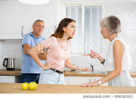 Elderly father tries to calm quarreled wife and daughter in home kitchen 99636383
