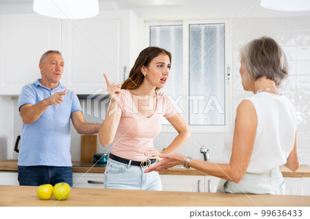 Elderly father tries to calm quarreled wife and daughter in home kitchen 99636433