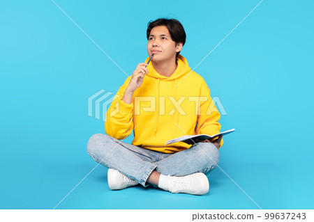 Thoughtful Korean Student Guy Holding Copybook Sitting Over Blue Background 99637243