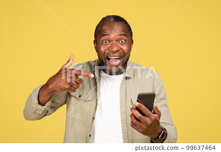 Glad excited shocked adult black guy in casual with open mouth pointing finger at smartphone, recommends app Glad excited shocked adult black guy in casual with open mouth pointing finger at smartphone, recommends app 99637464