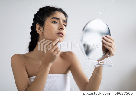 Worried Indian Female Holding Magnifying Mirror And Looking At Pimple On Chin 99637500