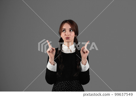 Pensive glad young girl student in gothic black clothes with pigtails shows fingers up to free space Pensive glad young girl student in gothic black clothes with pigtails shows fingers up to free space 99637549