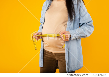 Young pregnant lady measuring her belly with tape to keep track of her fetus development, cropped, yellow background Young pregnant lady measuring her belly with tape to keep track of her fetus development, cropped, yellow background 99637603