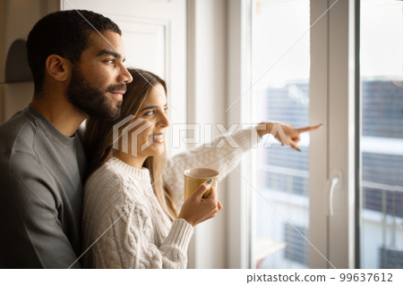 Happy young arab man hug european wife, lady pointing finger out the window and drink cup of tea 99637612