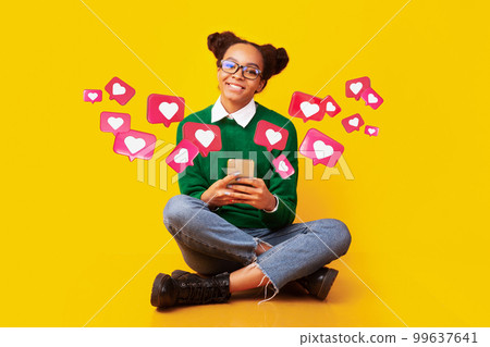 Happy teen african american female in glasses has romantic chat with hearts on phone, enjoys message 99637641