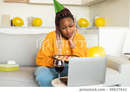 Sad single black woman in party hat celebrating birthday online, using laptop for video call, holding pie with candle 99637642
