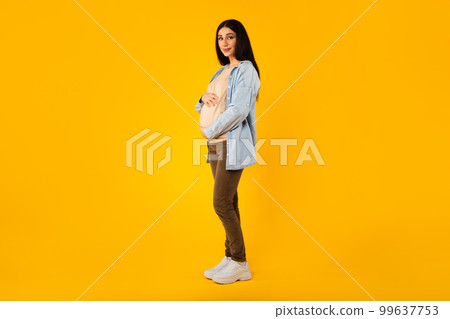 Young pregnant woman touching belly while posing on yellow studio background, full length shot, free space Young pregnant woman touching belly while posing on yellow studio background, full length shot, free space 99637753