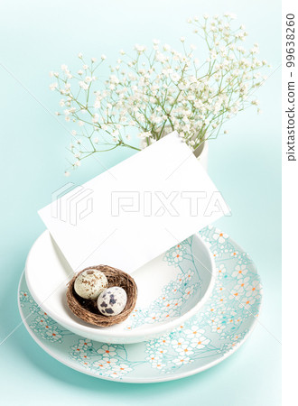 Easter card. Two quail eggs in nest, white empty note on plates with floral pattern on light blue. Easter card. Two quail eggs in nest, white empty note on plates with floral pattern on light blue. 99638260