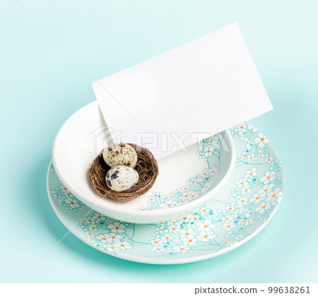 Table setting with Easter quail eggs in nest, white blank sheet on floral pattern plates on blue. 99638261