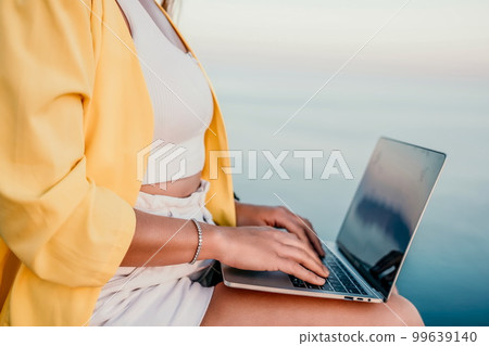 Digital nomad, Business woman working on laptop by the sea. Pretty lady typing on computer by the sea at sunset, makes a business transaction online from a distance. Freelance, remote work on vacation 99639140