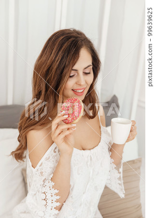 A brown-haired woman with long hair has breakfast in bed with a donut and a cup of coffee. The concept of a cozy home interior A brown-haired woman with long hair has breakfast in bed with a donut and a cup of coffee. The concept of a cozy home interior 99639625