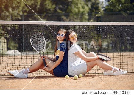 Two girls sitting on a tennis court and holding a rackets. Women wearing white and blue sport dresses and sunglasses. Girls standing together and posing for a photo. 99640334