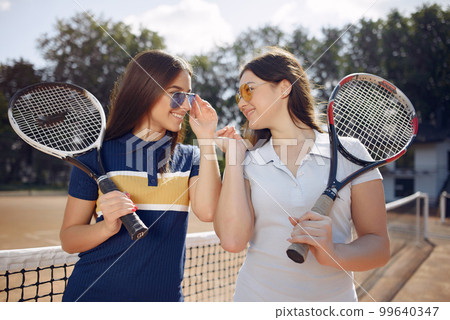 Two girls standing on a tennis court and holding a rackets. Women wearing white and blue sport dresses and sunglasses. Girls standing together and posing for a photo. 99640347