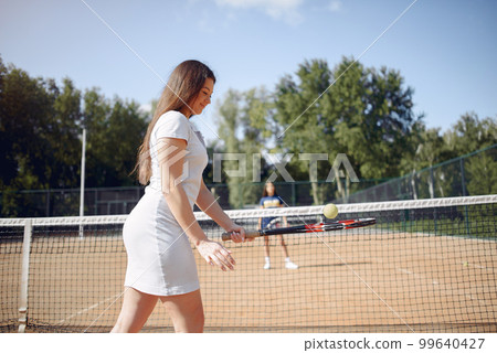 Two girls playing on a tennis court. Women wearing white and blue sport dresses. Focus on a girl in white dress. 99640427