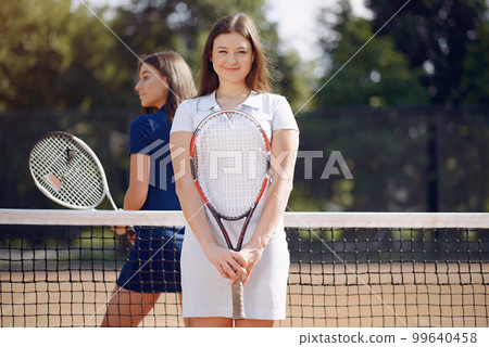 Two girls playing on a tennis court. Women wearing white and blue sport dresses. Focus on a girl in white dress, who looking at camera. Two girls playing on a tennis court. Women wearing white and blue sport dresses. Focus on a girl in white dress, who looking at camera. 99640458