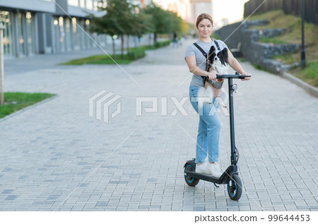 A woman rides an electric scooter with a dog in a backpack. Pappilion Spaniel Continental in a sling. A woman rides an electric scooter with a dog in a backpack. Pappilion Spaniel Continental in a sling. 99644453